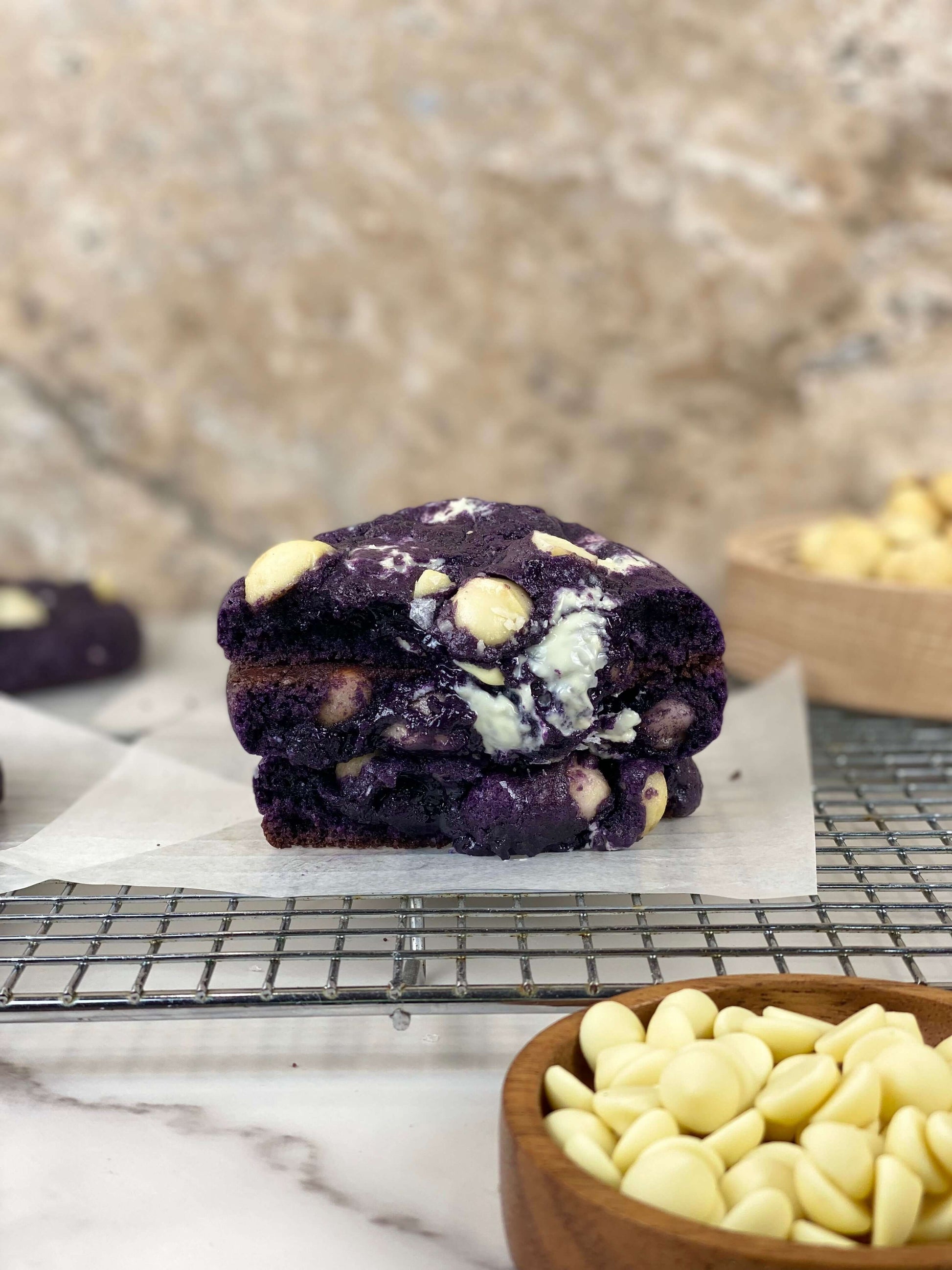Ube White Chocolate Macadamia Cookies stacked on a cooling rack with white chocolate chips in the foreground.